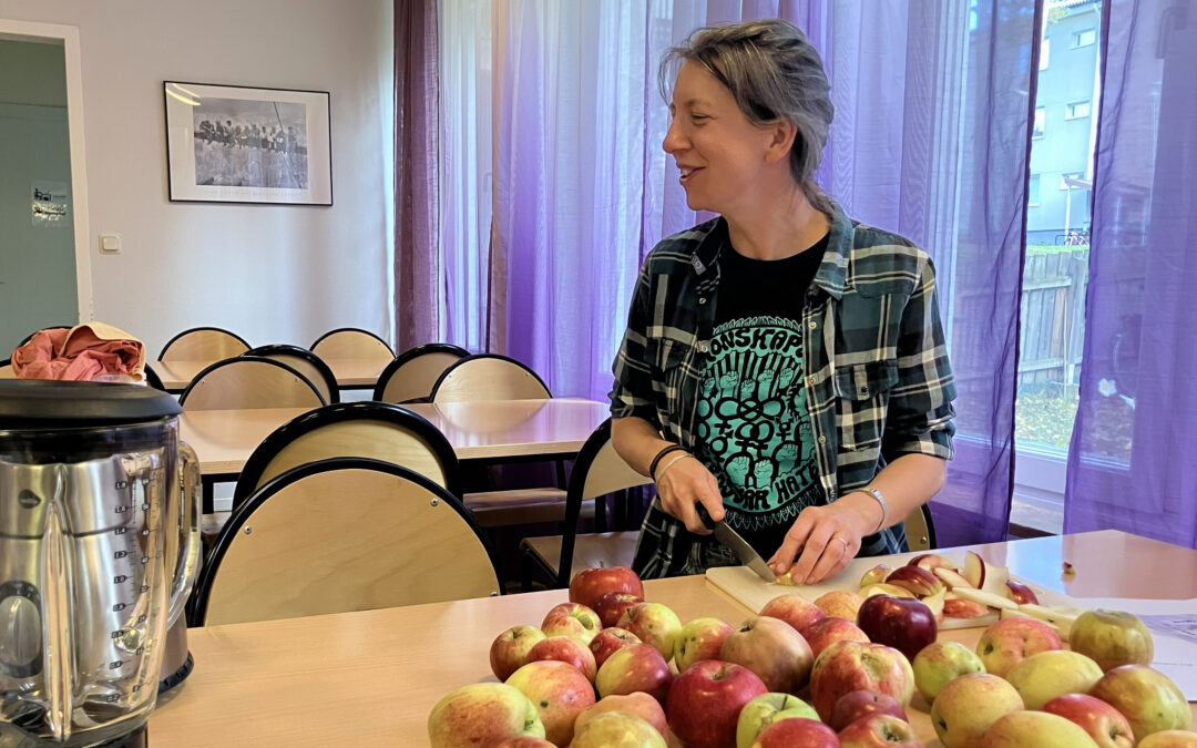 Kvinna sitter vi ett bord och skivar äpplen. Hon har rutig skjorta och svart t-shirt under med grönt tryck. Hon har hästsvans, cendréfärgat hår. Hon tittar åt höger och ser. Bakom henne fler bord och stolar.