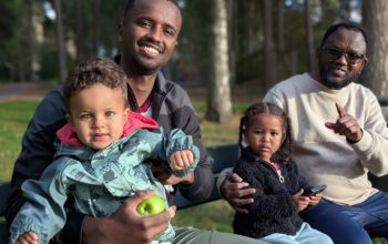 Två män med varsitt barn på en parkbänk. Mannen närmast kameran är svart med kort mörkt hår. Han har sin ett och ett halvt-åriga son i famnen och ler in i kameran. I handen håller han ett grönt äpple. Sonen är klädd i regnjacka och tittar in i kameran med ljusa ögon. mannen till höger i bild är också svart. Han har mörka glasögon och skägg. Han ler lätt och pekar mot kameran. Bredvid sig har han sin dotter, snart tre år. Hon har bruna flätor och ser lite skeptisk ut. I handen håller hon en mobiltelefon.