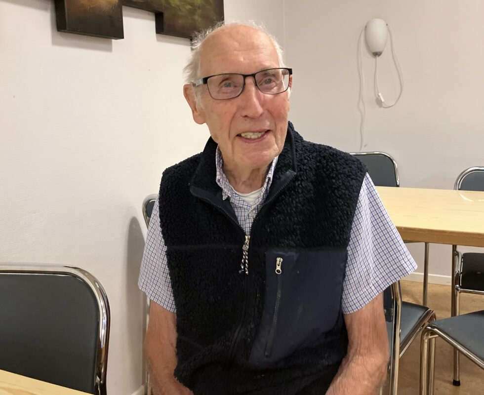 En 97-årig man sitter vid ett bord. Han tittar in i kameran och ler. Han hr vitt hår, kal hjässa, svart väst med kortärmad skjorta under.