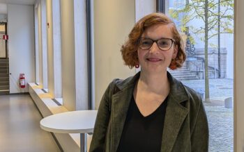 Halvkroppsporträtt på Claire Colomb, med vågigt brunrött hår i page, glasögon, grå kavaj, mörk topp, örhängen med röda ringar. Hon står i en korridor med vita väggar och en rad fönster, grått golv. Utanför fönstret närmast syns ett litet träd med gulgröna blad. Clarie Colomb är professor vid universitetet i Cambridge och har forskat på Airbnb och annan korttidsuthyrning.