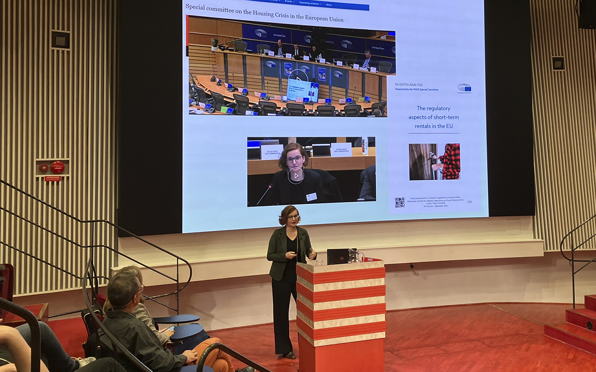 Claire Colomb står vid en pulpet och föreläser i amfiteaterformade Humanistiska teatern i Uppsala. Pulpeten är röd med ljusare ränder, golvet är rött. Till vänster ser man några åhörare. Bakom Claire Colomb en bild hur en presentation som visar EU-parlamentet, när hon talade där och rapporten hon skrev till EU-parlamentets tillfälliga utskott för bostadskrisen i Europeiska unionen. Professor Claire Colomb har forskat om Airbnb och annan korttidsuthyrning i tio år.