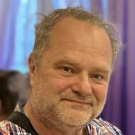 Porträttbild på en man i 50-årsåldern med rutig skjorta. Han har grått hår och lättare grå skäggväxt.