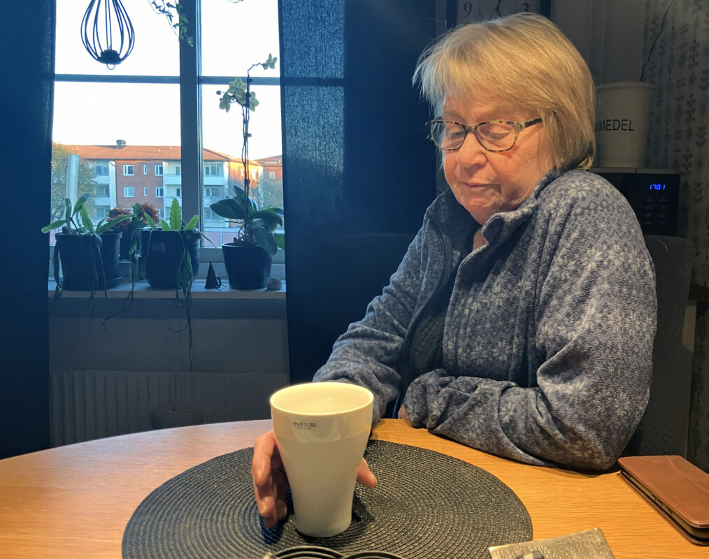 En kvinna i 70-årsåldern sitter vid ett köksbord. Hon håller i en kaffekopp och tittar ner mot bordet. Hon har kort kort, glasögon och grå tröja. I bakgrunden ett köksfönster med mörkblå gardiner och krukväxter. 