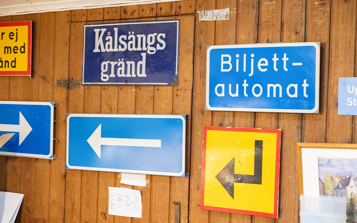 Vägskyltar av olika slag på en vägg av lodräta träplankor. På skyltarna står det Kålsängsgränd och Biljettautomat. På två stycken är det pilar som pekar till vänster.