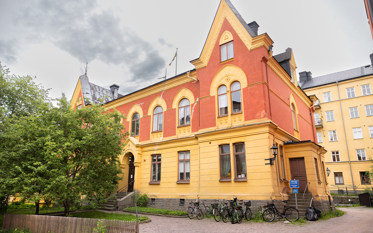 En gul stenbyggnad från 1800-talet i rött och gult. Framför ingången är ett grönskande träd. Utanför står några cyklar parkerade,