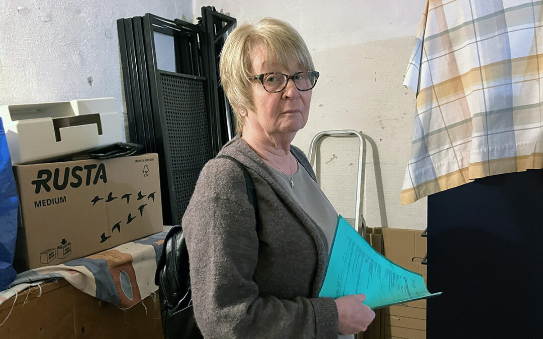 Kvinna i 70-årsåldern står i ett källarförråd. Hon har kort blont hår, glasögon och tittar allvarligt in i kameran. I handen håller hon en grön mapp. I bakgrunden kartonger.