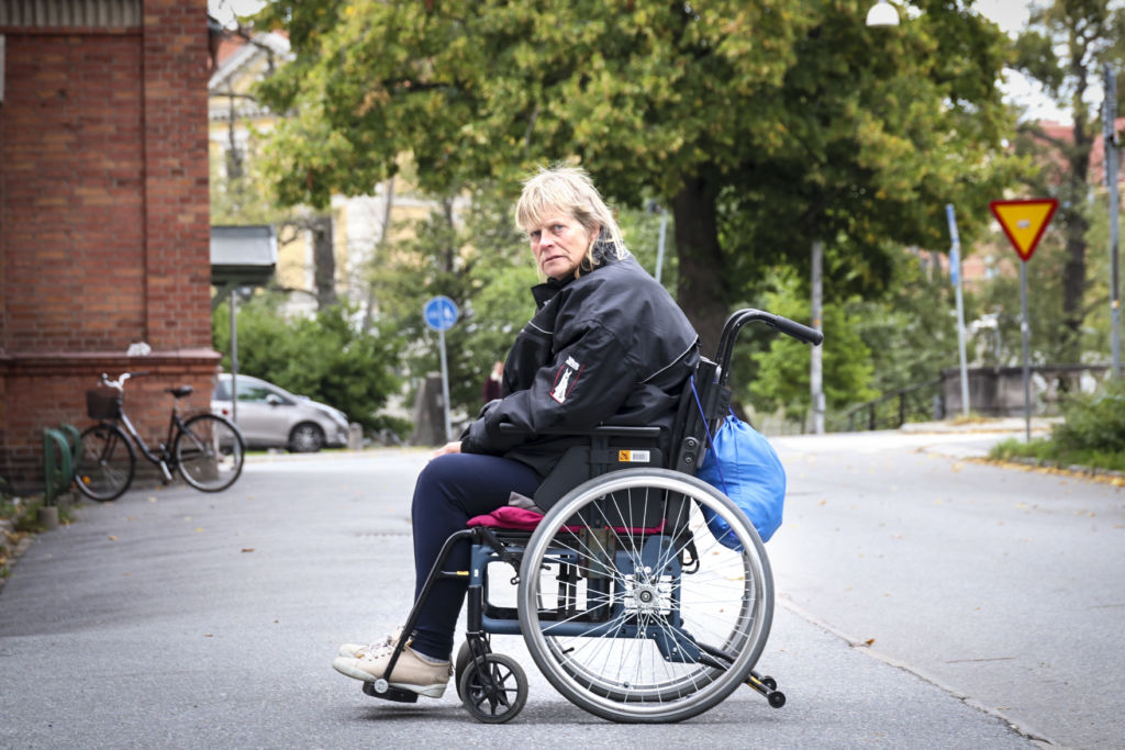 En kvinna sitter i en rullstol på en trottoar. Hon är ensam, tittar in i kameran. En blå påse hänger bak på rullstolen.