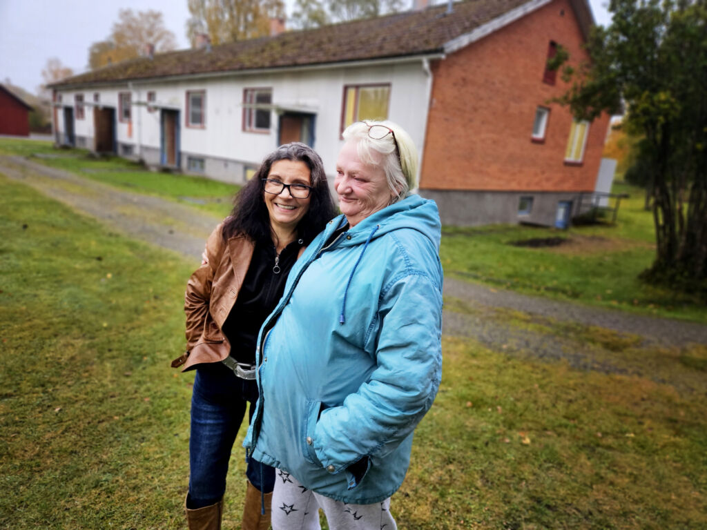 Två kvinnor står intill varandra en grå höstdag. Den ena i mörkt hår och brun skinnjacka. Den andra med ljust hår och en turkos jacka.