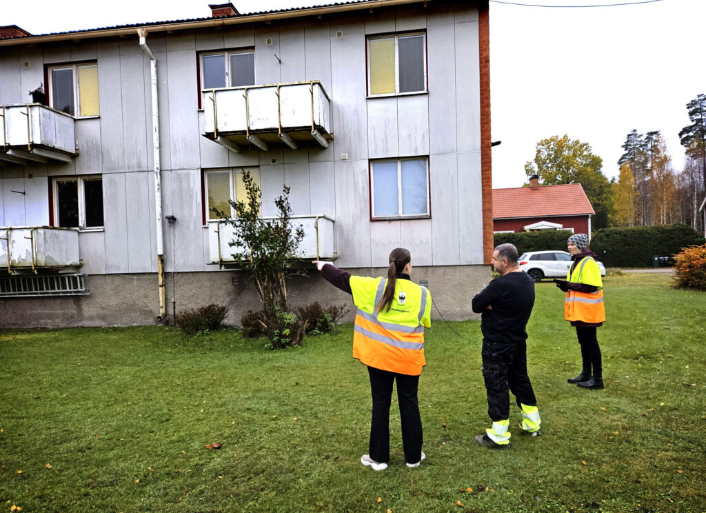 Två inspektörer i orange västar står med ryggarna mot kameran och inspekterar ett grått hyreshus tillsammans med en man i svarta kläder.