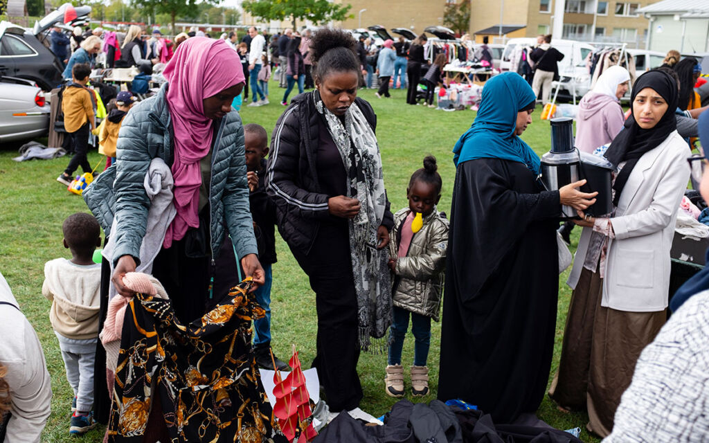 Flera kvinnor som tillsammans med barn tittar på olika saker på en loppmarknad. Kvinnorna ser ut att ha sitt ursprung i Afrika och Mellanöstern och håller i kläder och en köksapparat. I bildkanten skymtar säljaren.